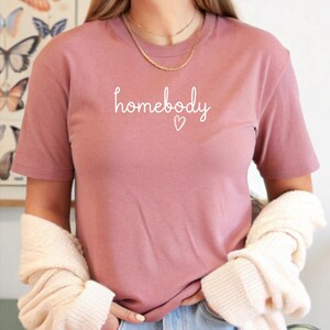 Puede incluir: Una camiseta de color rosa polvoriento con la palabra "homebody" en escritura blanca, acompa&ntilde;ada de un peque&ntilde;o coraz&oacute;n. La camiseta de manga corta parece estar hecha de un material suave. La modelo lleva vaqueros azules y un su&eacute;ter color crema.