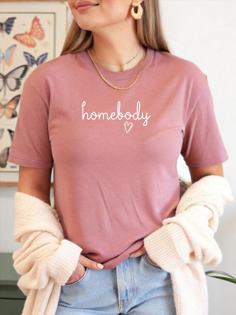 Puede incluir: Una camiseta de color rosa polvoriento con la palabra "homebody" en escritura blanca, acompa&ntilde;ada de un peque&ntilde;o coraz&oacute;n. La camiseta de manga corta parece estar hecha de un material suave. La modelo lleva vaqueros azules y un su&eacute;ter color crema.