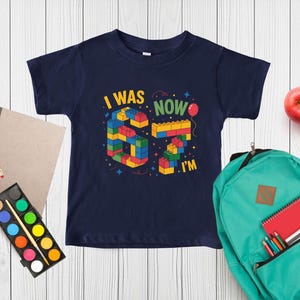 May include: A navy blue short-sleeve t-shirt featuring a graphic of the number 67 constructed from colorful interlocking plastic building blocks. The text reads I WAS 67 NOW I'M with a small red balloon and scattered star shapes in the design.