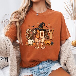 May include: A burnt orange short-sleeve t-shirt featuring a graphic print with the word SPOOKY in varied patterns, a black witch hat, a large orange and white checkered bow, pumpkins, autumn leaves, a small ghost, and bats.