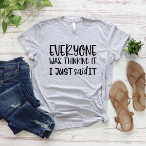 May include: A light gray t-shirt with the text "EVERYONE WAS THINKING IT I JUST SAID IT" in black. The shirt is paired with blue jeans and brown sandals. Green plants are also visible.