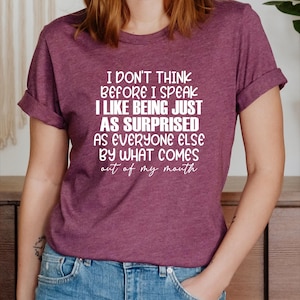 May include: Burgundy t-shirt with the text "I don't think before I speak, I like being just as surprised as everyone else by what comes out of my mouth" in white lettering. The shirt is paired with blue jeans.