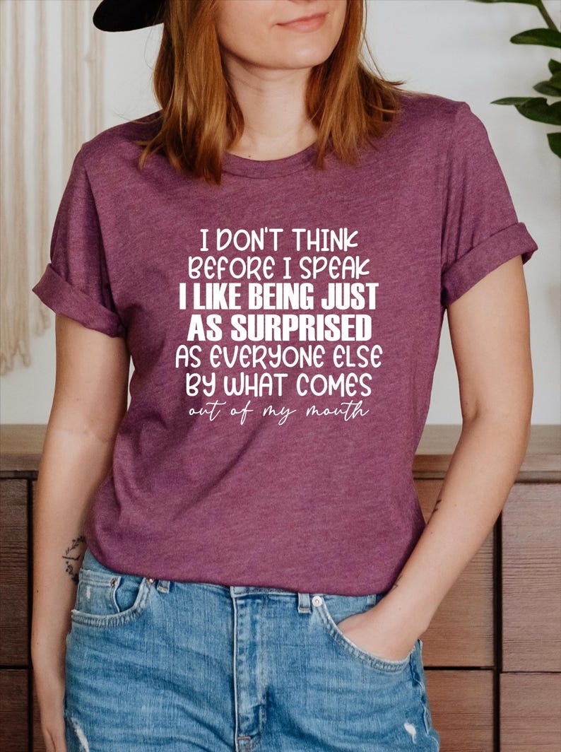 May include: Burgundy t-shirt with the text "I don't think before I speak, I like being just as surprised as everyone else by what comes out of my mouth" in white lettering. The shirt is paired with blue jeans.