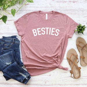 May include: A dusty rose t-shirt with the word "BESTIES" printed in white on the front. The t-shirt is laid flat on a white wooden surface with a pair of blue jeans and brown sandals.