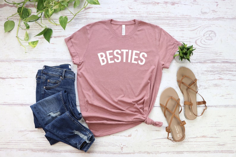 May include: A dusty rose t-shirt with the word "BESTIES" printed in white on the front. The t-shirt is laid flat on a white wooden surface with a pair of blue jeans and brown sandals.