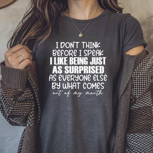 May include: Dark gray t-shirt with the text "I DON'T THINK BEFORE I SPEAK I LIKE BEING JUST AS SURPRISED AS EVERYONE ELSE BY WHAT COMES out of my mouth" in white. The person is wearing a necklace and a patterned jacket.
