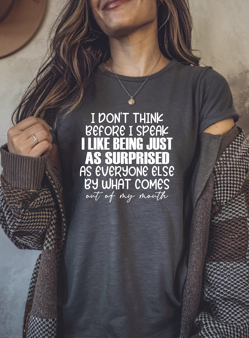 May include: Dark gray t-shirt with the text "I DON'T THINK BEFORE I SPEAK I LIKE BEING JUST AS SURPRISED AS EVERYONE ELSE BY WHAT COMES out of my mouth" in white. The person is wearing a necklace and a patterned jacket.