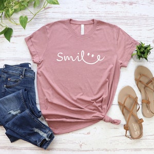 May include: A dusty rose t-shirt with the text "Smile" in white lettering. The shirt is knotted at the bottom and is laid out on a white wooden surface with a pair of brown sandals and blue jeans.