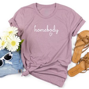 Puede incluir: Una camiseta de color malva con la palabra "homebody" en escritura blanca y un peque&ntilde;o coraz&oacute;n. La camiseta est&aacute; combinada con pantalones cortos de mezclilla, gafas de sol, sandalias y flores blancas.