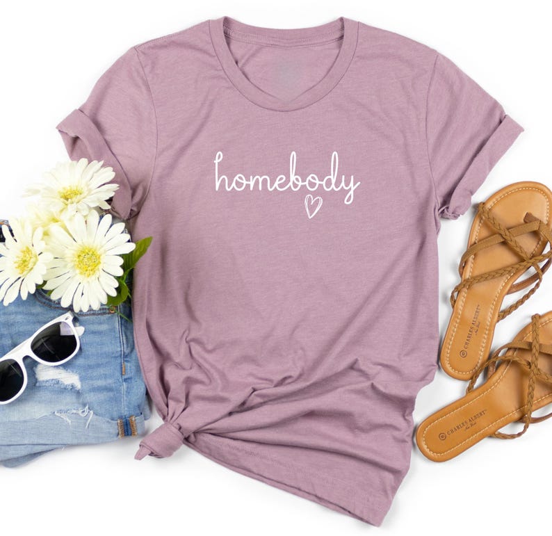 Puede incluir: Una camiseta de color malva con la palabra "homebody" en escritura blanca y un peque&ntilde;o coraz&oacute;n. La camiseta est&aacute; combinada con pantalones cortos de mezclilla, gafas de sol, sandalias y flores blancas.