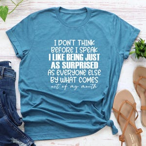 May include: Teal blue t-shirt with the text "I DON'T THINK BEFORE I SPEAK I LIKE BEING JUST AS SURPRISED AS EVERYONE ELSE BY WHAT COMES out of my mouth" in white. The shirt is knotted at the bottom.