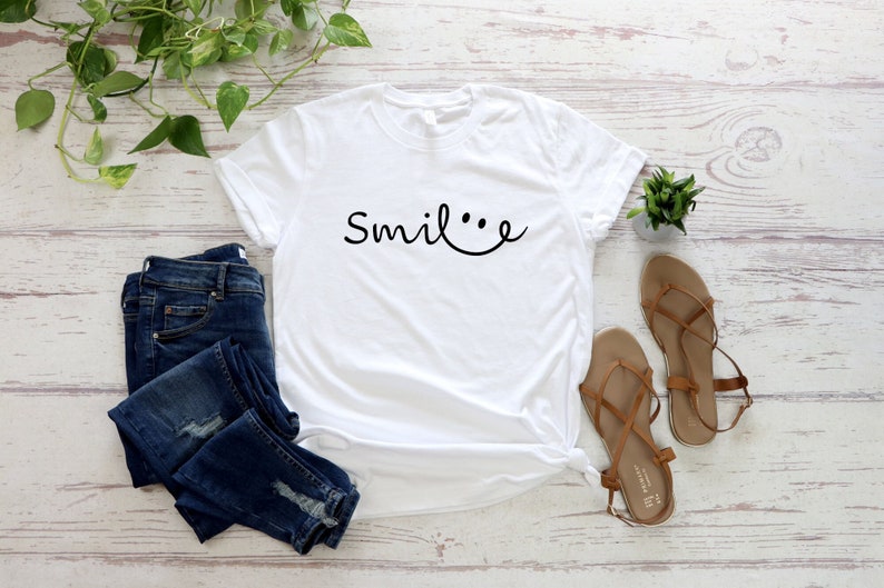 May include: White t-shirt with the text "Smile..." in black cursive font, featuring a smiley face with two dots for eyes. The shirt is laid flat on a white wooden surface with a pair of brown sandals, blue jeans, and a small potted plant.