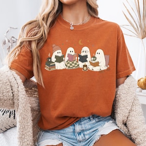 May include: A burnt orange short-sleeve t-shirt featuring a graphic print of four white ghosts sitting together. The ghosts are reading books, wearing a beanie and glasses, and drinking from mugs. The design includes small stars and a crescent moon.