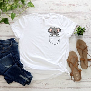 May include: White t-shirt with a front pocket featuring a cartoon koala bear. The shirt is laid flat on a white wooden surface with a pair of brown sandals and blue jeans.