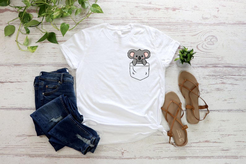 May include: White t-shirt with a front pocket featuring a cartoon koala bear. The shirt is laid flat on a white wooden surface with a pair of brown sandals and blue jeans.