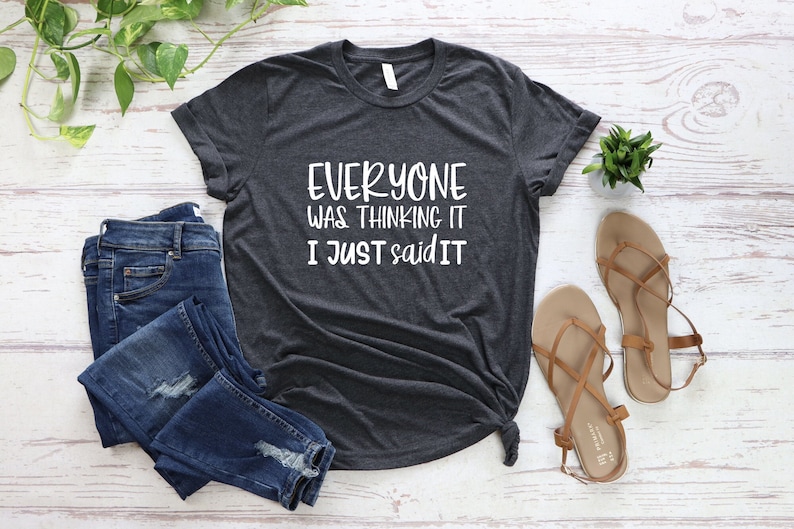 May include: Dark gray t-shirt with the text "EVERYONE WAS THINKING IT I JUST SAID IT" in white. The shirt is paired with blue jeans and brown sandals. A small green plant is also visible.