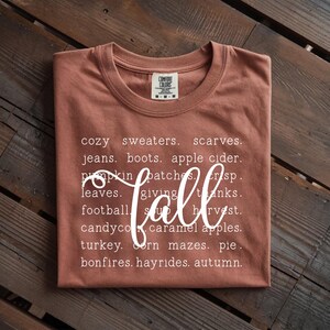 May include: A terracotta-colored short-sleeve t-shirt featuring white text listing fall-themed words including cozy sweaters, scarves, jeans, boots, apple cider, pumpkin patches, crisp, leaves, giving, thanks, football, soup, harvest, candy corn, caramel apples, turkey, corn mazes, pie, bonfires, hayrides, autumn, and the word fall in a large script font.
