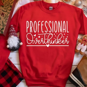 A red sweatshirt with white text that says "PROFESSIONAL OVERTHINKER" is displayed on a table surrounded by various holiday-themed items, including a gnome, a gingerbread man, and a pair of boots.