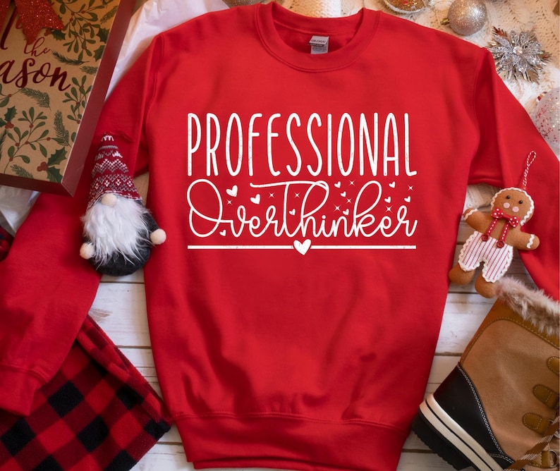 A red sweatshirt with white text that says "PROFESSIONAL OVERTHINKER" is displayed on a table surrounded by various holiday-themed items, including a gnome, a gingerbread man, and a pair of boots.