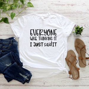 May include: White t-shirt with the text "EVERYONE WAS THINKING IT I JUST SAID IT" in black. The shirt is paired with blue denim jeans and brown sandals. A small green plant and decorative greenery are also in the image.
