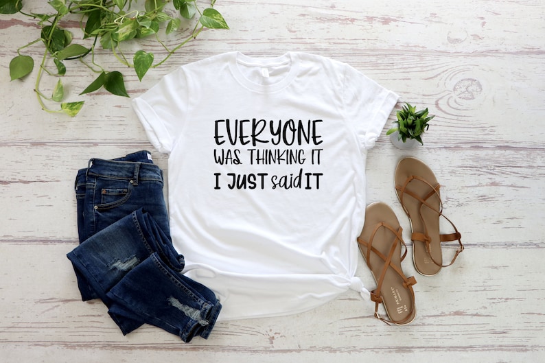 May include: White t-shirt with the text "EVERYONE WAS THINKING IT I JUST SAID IT" in black. The shirt is paired with blue denim jeans and brown sandals. A small green plant and decorative greenery are also in the image.