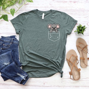 May include: A green t-shirt with a pocket featuring a cartoon koala. The koala is peeking out of the pocket with its arms crossed. The t-shirt is laid out on a white wooden surface with a pair of brown sandals and a pair of blue jeans.