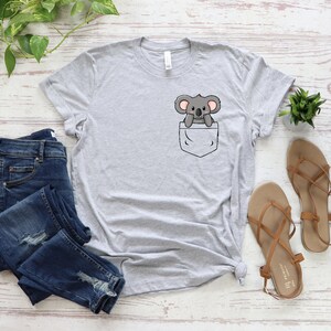 May include: A gray t-shirt with a pocket featuring a cartoon koala peeking out. The shirt is laid flat on a white wooden surface with a pair of brown sandals and blue jeans.