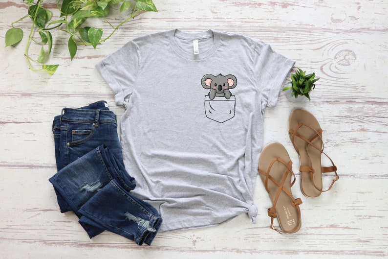 May include: A gray t-shirt with a pocket featuring a cartoon koala peeking out. The shirt is laid flat on a white wooden surface with a pair of brown sandals and blue jeans.