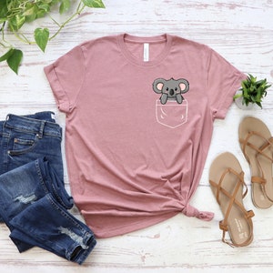 May include: A pink t-shirt with a front pocket featuring a cartoon koala. The koala is gray and has a white belly. The shirt is made of a soft, comfortable material.