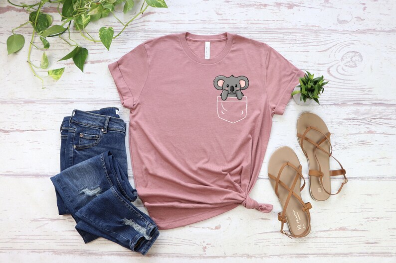 May include: A pink t-shirt with a front pocket featuring a cartoon koala. The koala is gray and has a white belly. The shirt is made of a soft, comfortable material.