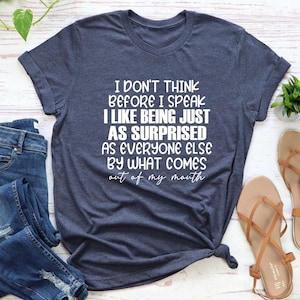 May include: A navy blue t-shirt with the text "I DON'T THINK BEFORE I SPEAK I LIKE BEING JUST AS SURPRISED AS EVERYONE ELSE BY WHAT COMES out of my mouth" in white. The shirt is styled with jeans and sandals.