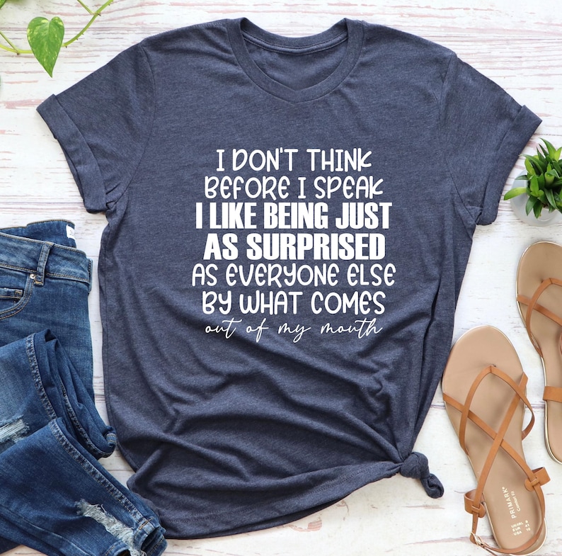 May include: A navy blue t-shirt with the text "I DON'T THINK BEFORE I SPEAK I LIKE BEING JUST AS SURPRISED AS EVERYONE ELSE BY WHAT COMES out of my mouth" in white. The shirt is styled with jeans and sandals.