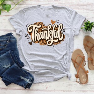 May include: A light gray short-sleeve t-shirt featuring the word Thankful in a cream-colored script font over a brown leopard print background, accompanied by small heart and leaf illustrations, paired with dark blue distressed denim jeans.