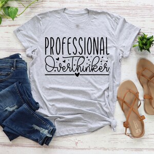 a casual outfit consisting of a gray t-shirt with the text "Professional Overthinker" printed on it, a pair of blue jeans, and a pair of brown sandals. The outfit is laid out on a white surface, with a potted plant and a small plant visible in the background.