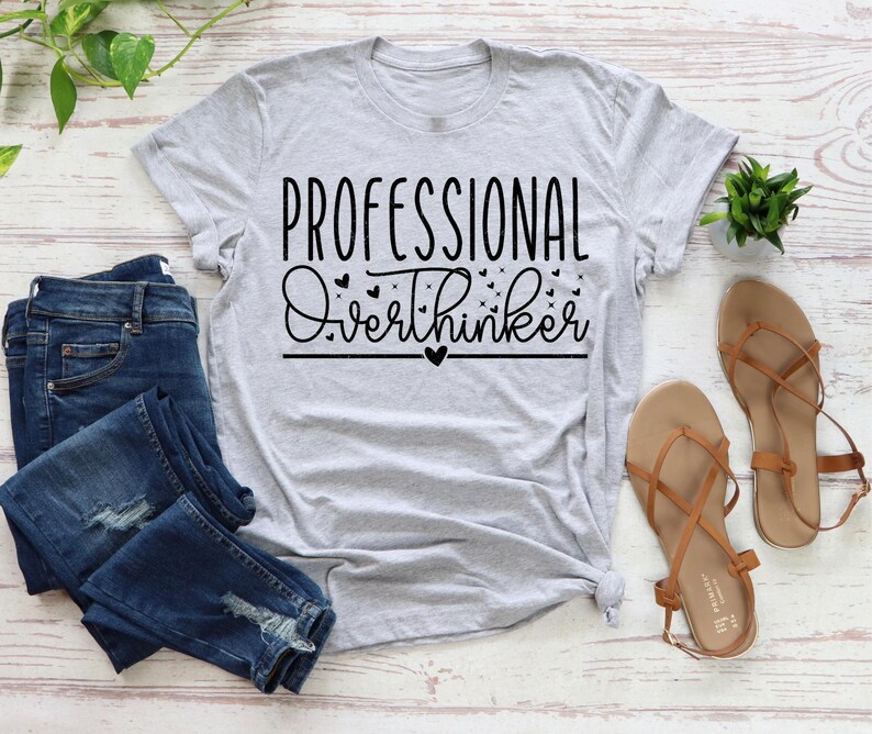 a casual outfit consisting of a gray t-shirt with the text "Professional Overthinker" printed on it, a pair of blue jeans, and a pair of brown sandals. The outfit is laid out on a white surface, with a potted plant and a small plant visible in the background.