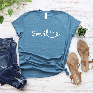 May include: A blue t-shirt with the text "Smile..." in white lettering. The shirt is styled with blue jeans and brown sandals.