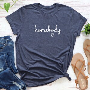 Puede incluir: Una camiseta azul marino con la palabra "homebody" en escritura blanca y un peque&ntilde;o coraz&oacute;n. La camiseta tiene las mangas enrolladas y un nudo en la parte inferior. Tambi&eacute;n se ven unos vaqueros azules y unas sandalias marrones.