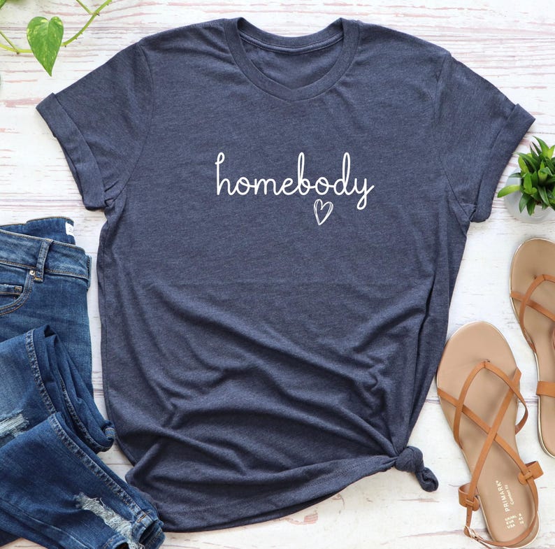 Puede incluir: Una camiseta azul marino con la palabra "homebody" en escritura blanca y un peque&ntilde;o coraz&oacute;n. La camiseta tiene las mangas enrolladas y un nudo en la parte inferior. Tambi&eacute;n se ven unos vaqueros azules y unas sandalias marrones.