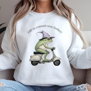 May include: White sweatshirt featuring a green frog wearing a purple wizard hat and riding a white scooter. The text "Off to commit tom foolery" is arched above the frog. The sweatshirt is a casual, comfortable style.