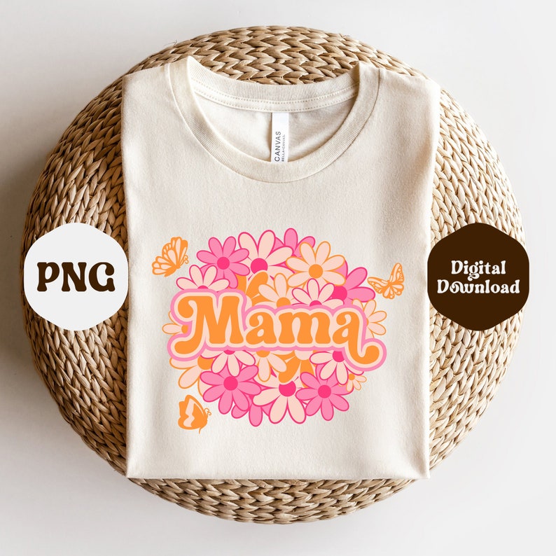 Puede incluir: Una camiseta blanca con un dise&ntilde;o gr&aacute;fico de la palabra "Mama" en letras naranja y rosa rodeada de flores y mariposas rosas y naranjas.