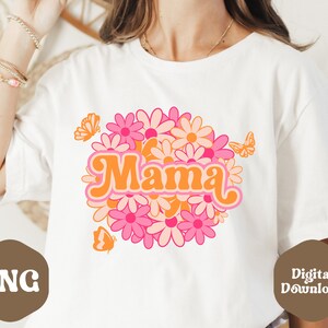 Puede incluir: Camiseta blanca con un dise&ntilde;o gr&aacute;fico retro que presenta la palabra "Mama" rodeada de margaritas rosas y naranjas y mariposas.