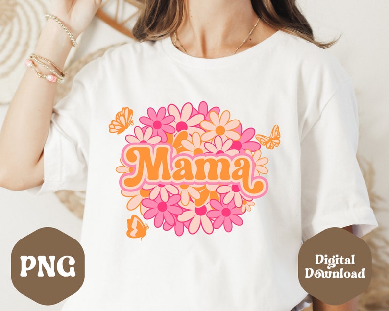 Puede incluir: Camiseta blanca con un dise&ntilde;o gr&aacute;fico retro que presenta la palabra "Mama" rodeada de margaritas rosas y naranjas y mariposas.