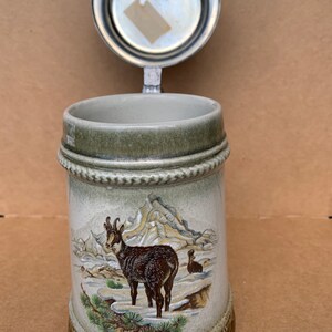 May include: A ceramic beer stein with a pewter lid. The stein features a brown and white mountain goat in a snowy mountain scene. The lid has a silver finish.