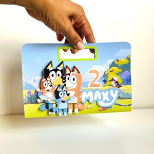May include: A colourful birthday card featuring the characters from the Bluey cartoon. The card is decorated with a blue, green and yellow background and features the characters Bluey, Bingo and Bandit. The card also includes the number "2" and the name "Maxy".