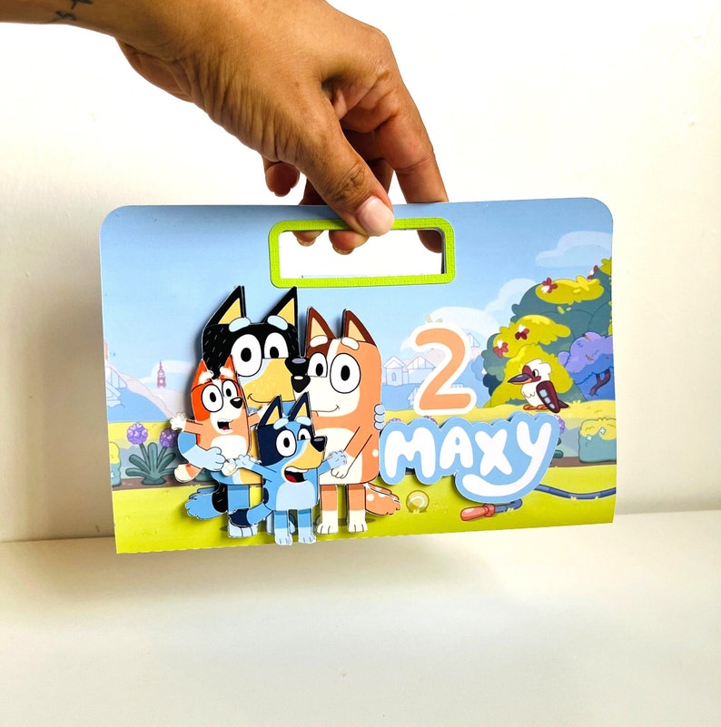 May include: A colourful birthday card featuring the characters from the Bluey cartoon. The card is decorated with a blue, green and yellow background and features the characters Bluey, Bingo and Bandit. The card also includes the number "2" and the name "Maxy".