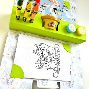 May include: A green and white cardboard activity set featuring a colouring page of two cartoon dogs, crayons, Play-Doh, and two smiley face stamps.