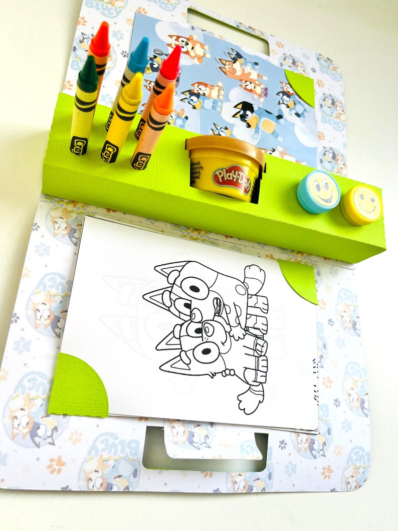 May include: A green and white cardboard activity set featuring a colouring page of two cartoon dogs, crayons, Play-Doh, and two smiley face stamps.