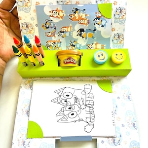 May include: A blue and white patterned cardboard box with a green handle. The box is open and contains a colouring book, crayons, Play-Doh, and two smiley face stamps. The colouring book features a black and white illustration of two cartoon dogs.