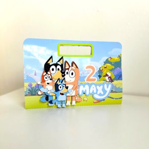 May include: A colourful birthday card featuring the characters from the cartoon show Bluey. The card is blue and green with a white border. The characters are a blue dog, a red dog, a brown dog, and an orange dog. The card says "Maxy" and "2" on it.