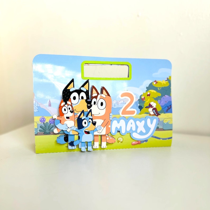 May include: A colourful birthday card featuring the characters from the cartoon show Bluey. The card is blue and green with a white border. The characters are a blue dog, a red dog, a brown dog, and an orange dog. The card says "Maxy" and "2" on it.